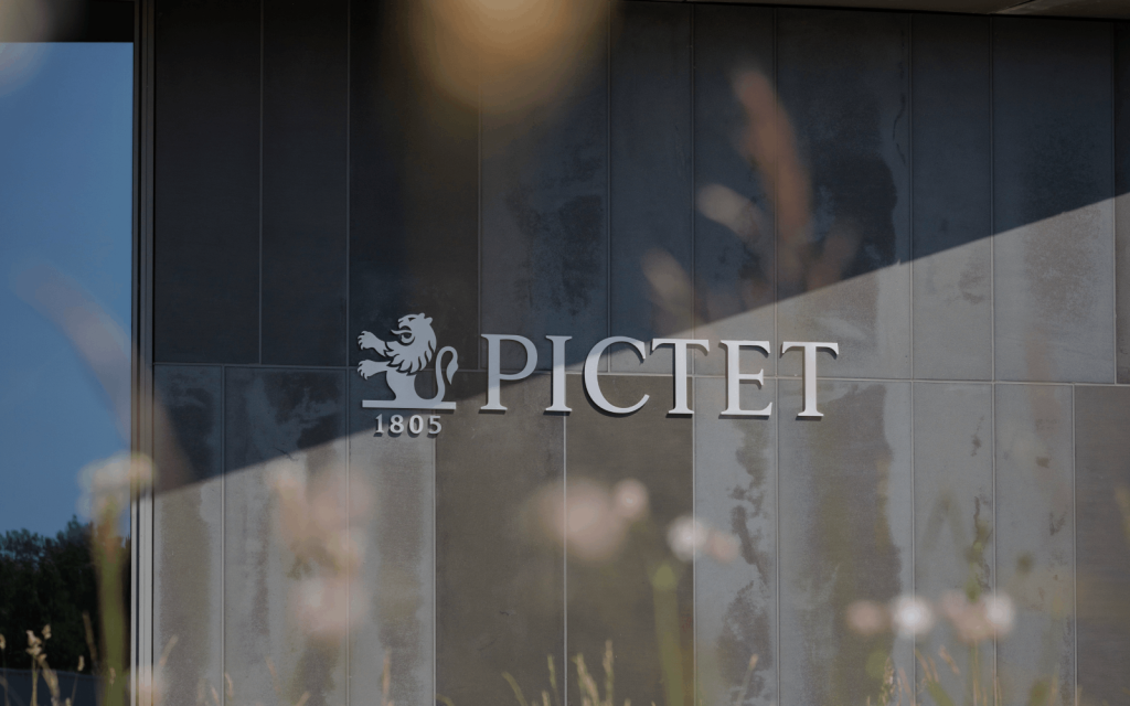 Pictet Asset Management logo featuring a lion emblem and the year 1805, displayed on a glass window with a blurred urban background.
