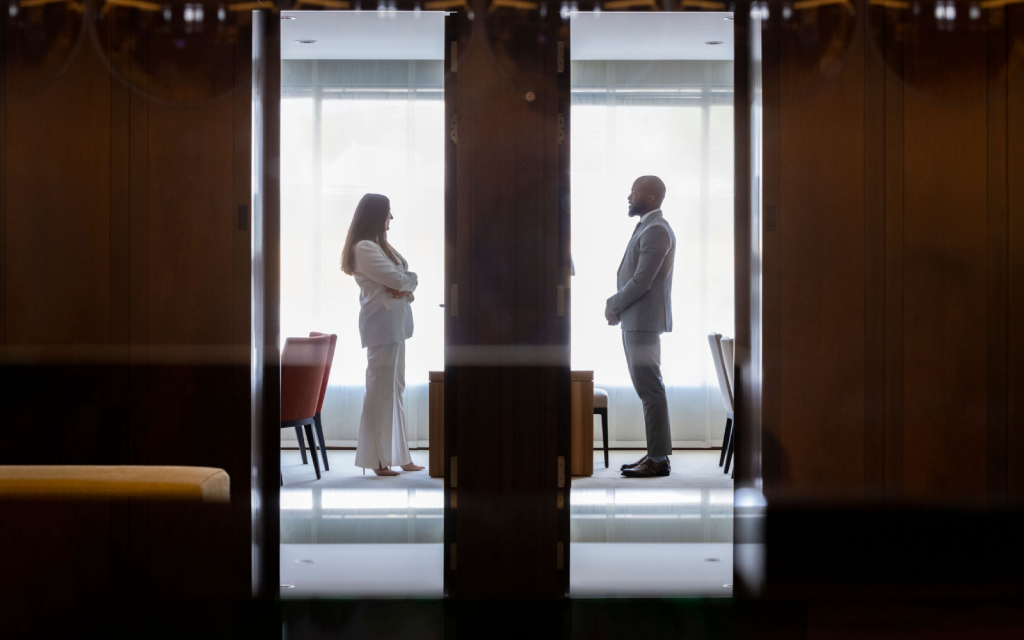 Two professionals in formal attire standing face-to-face in a modern office setting, separated by a glass partition, symbolizing a discussion or negotiation.