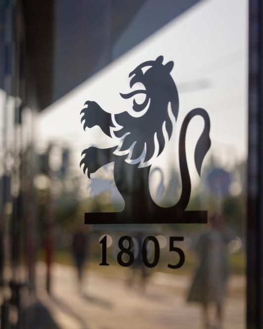 Pictet Asset Management logo featuring a lion emblem and the year 1805, displayed on a glass window with a blurred urban background.