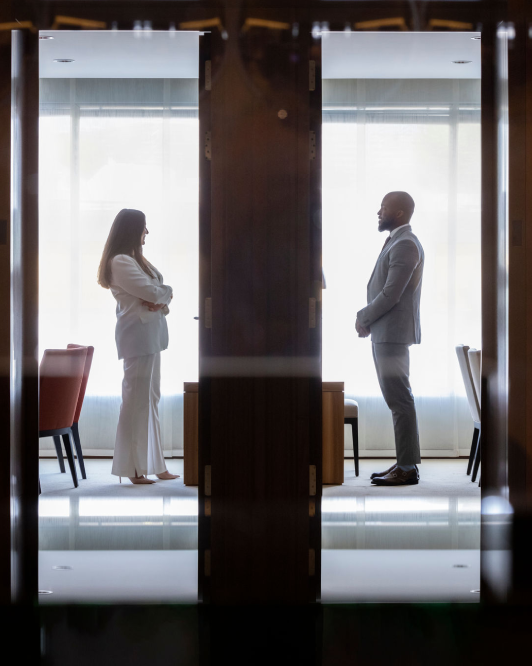 Two professionals in formal attire standing face-to-face in a modern office setting, separated by a glass partition, symbolizing a discussion or negotiation.